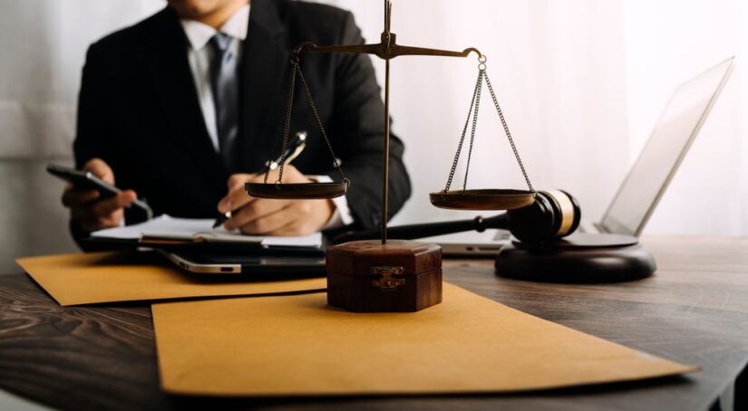 Lawyer Filling Out Paperwork With Scales Of Law On His Table